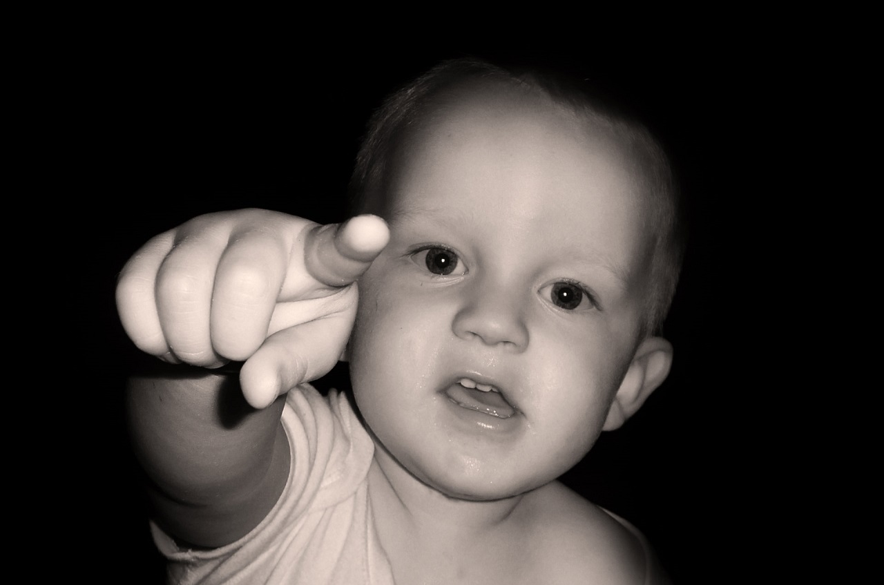 A baby, not yet a year old, extends one finger to point straight at the camera against a dark background