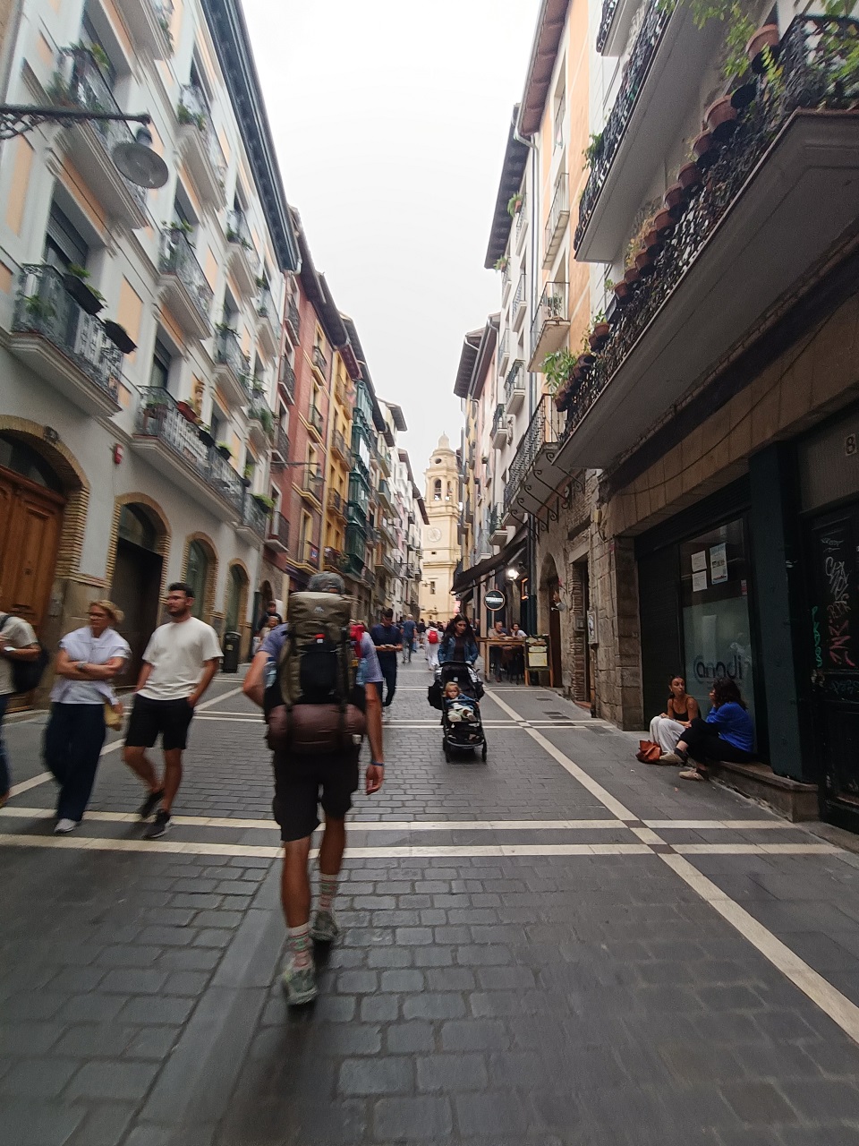 A walker with a large backpack, seen from behind, on a cobbled street in Pamplona&rsquo;s old town, heading toward the cathedral tower