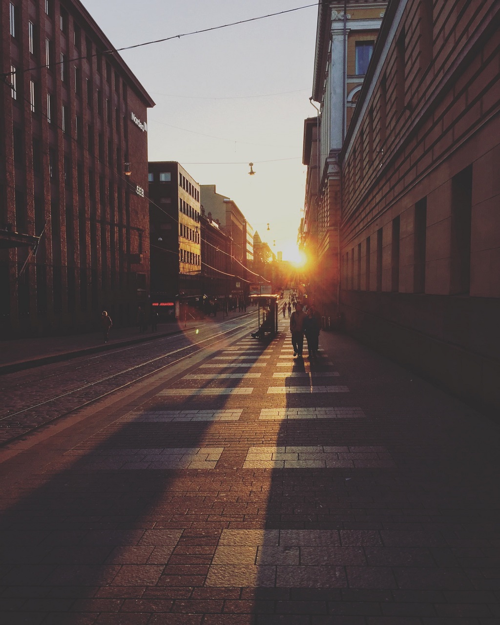 Two people walking along a sunlit city street at sunset, with tram tracks beside the pavement